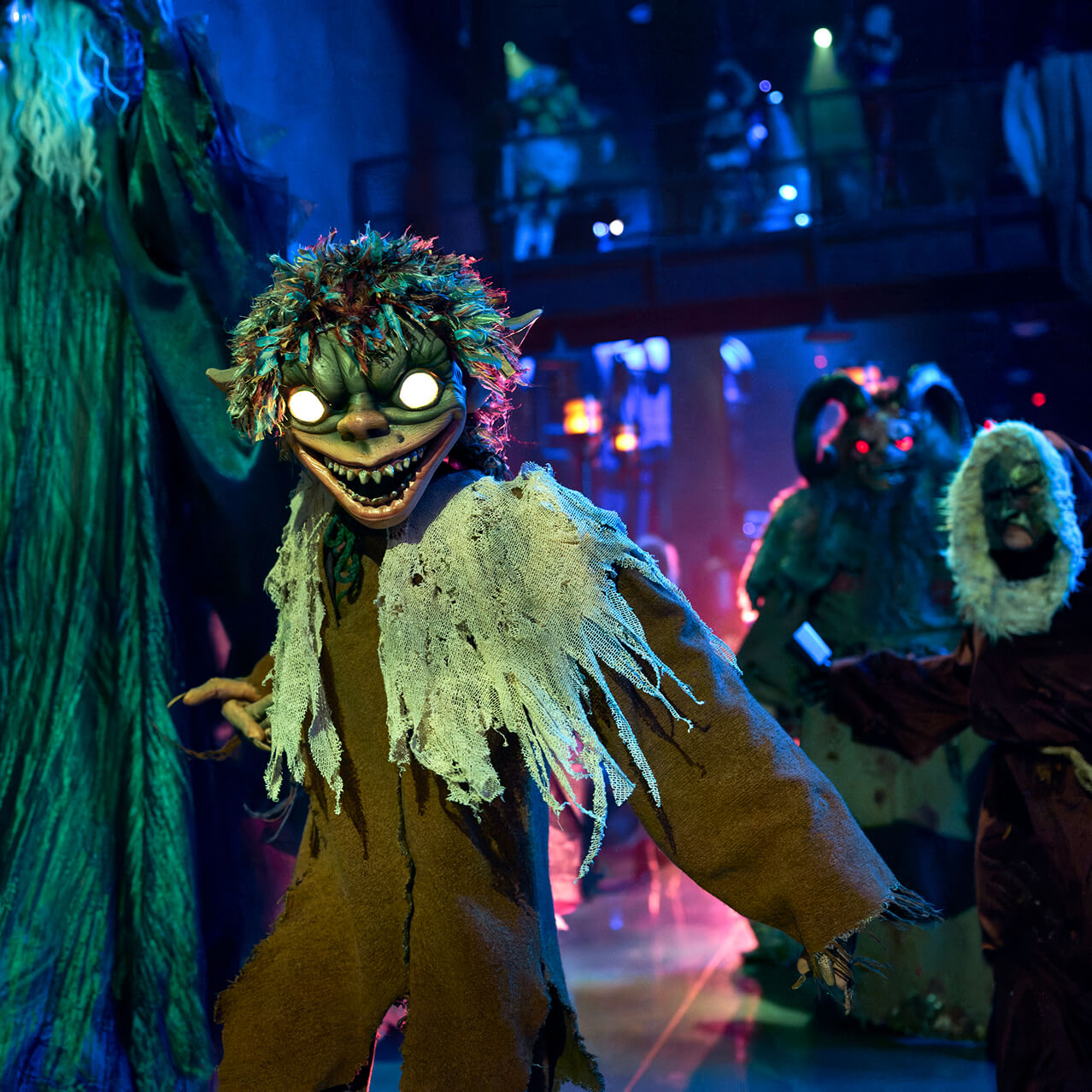 A procession of monsters walk forward as Kallikantzaroi grins with glowing yellow eyes and looks at the camera.