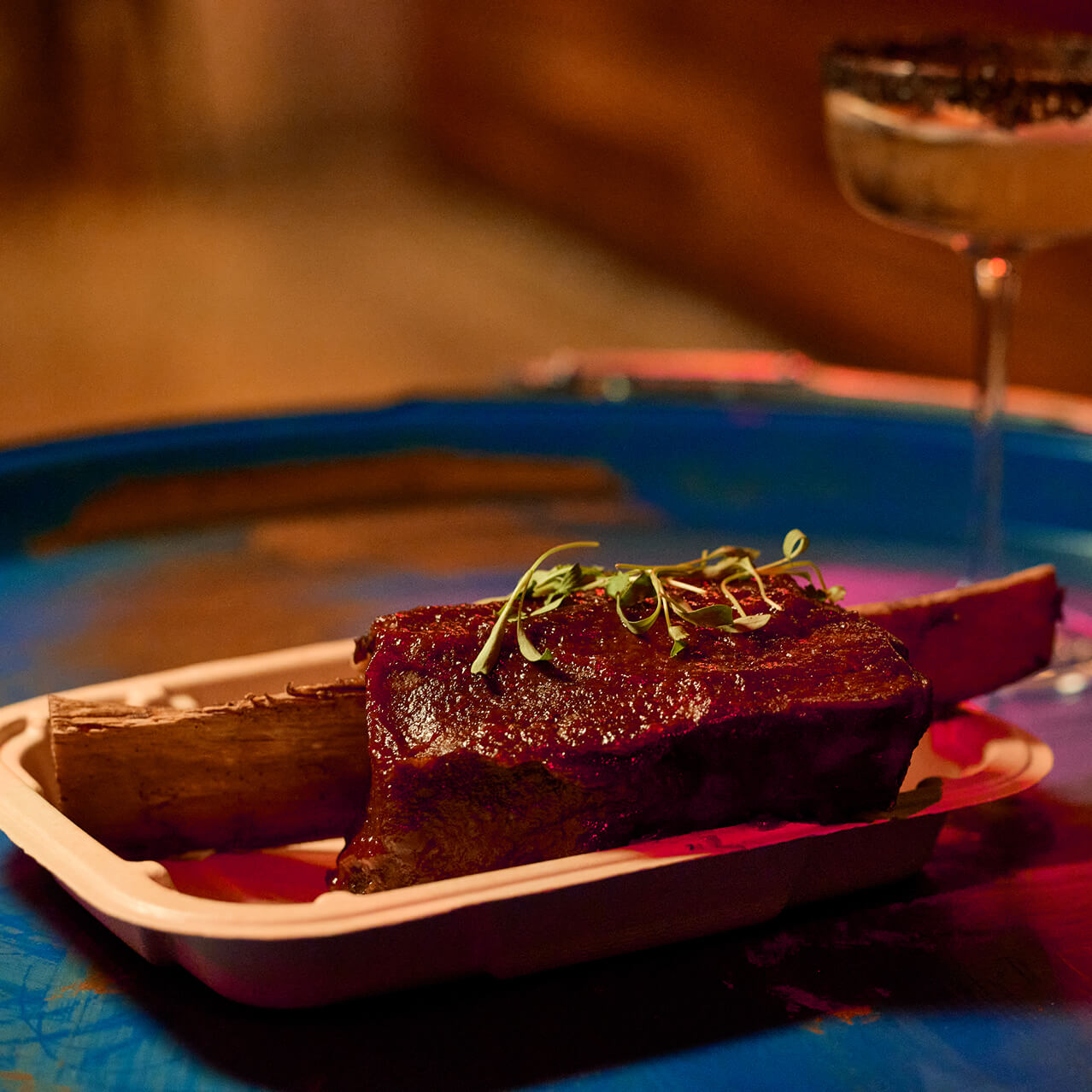 Large glazed beef rib with bone on tray, cocktail in the background