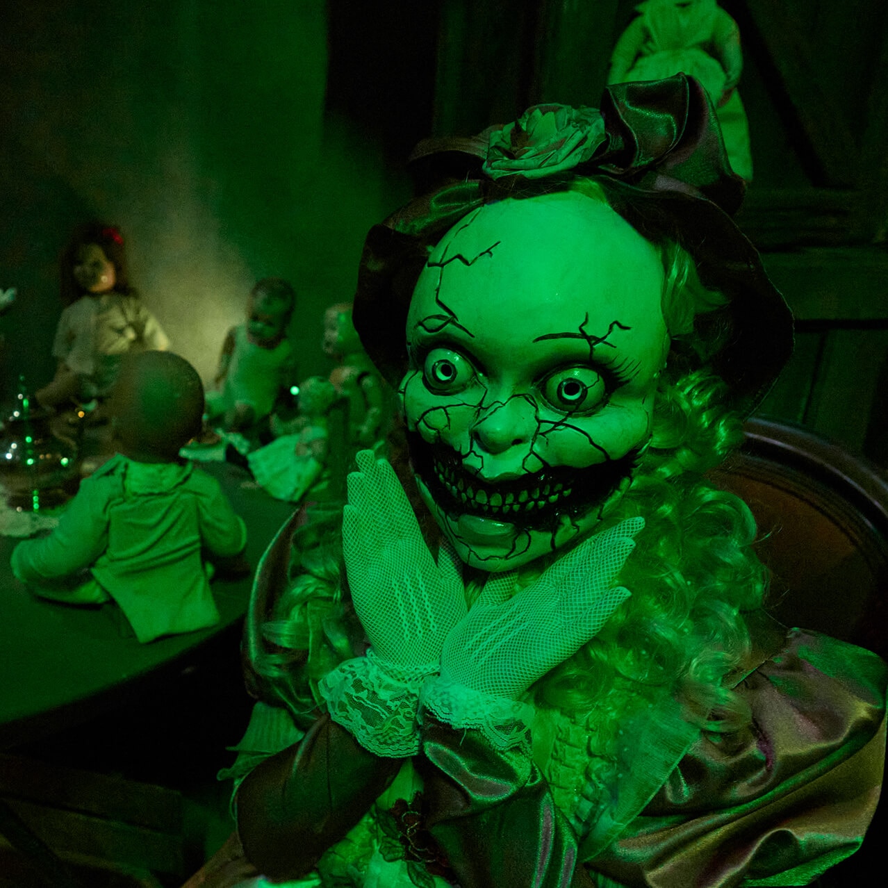 Creepy cracked doll figure with wide eyes in a room filled with dolls under green lighting.