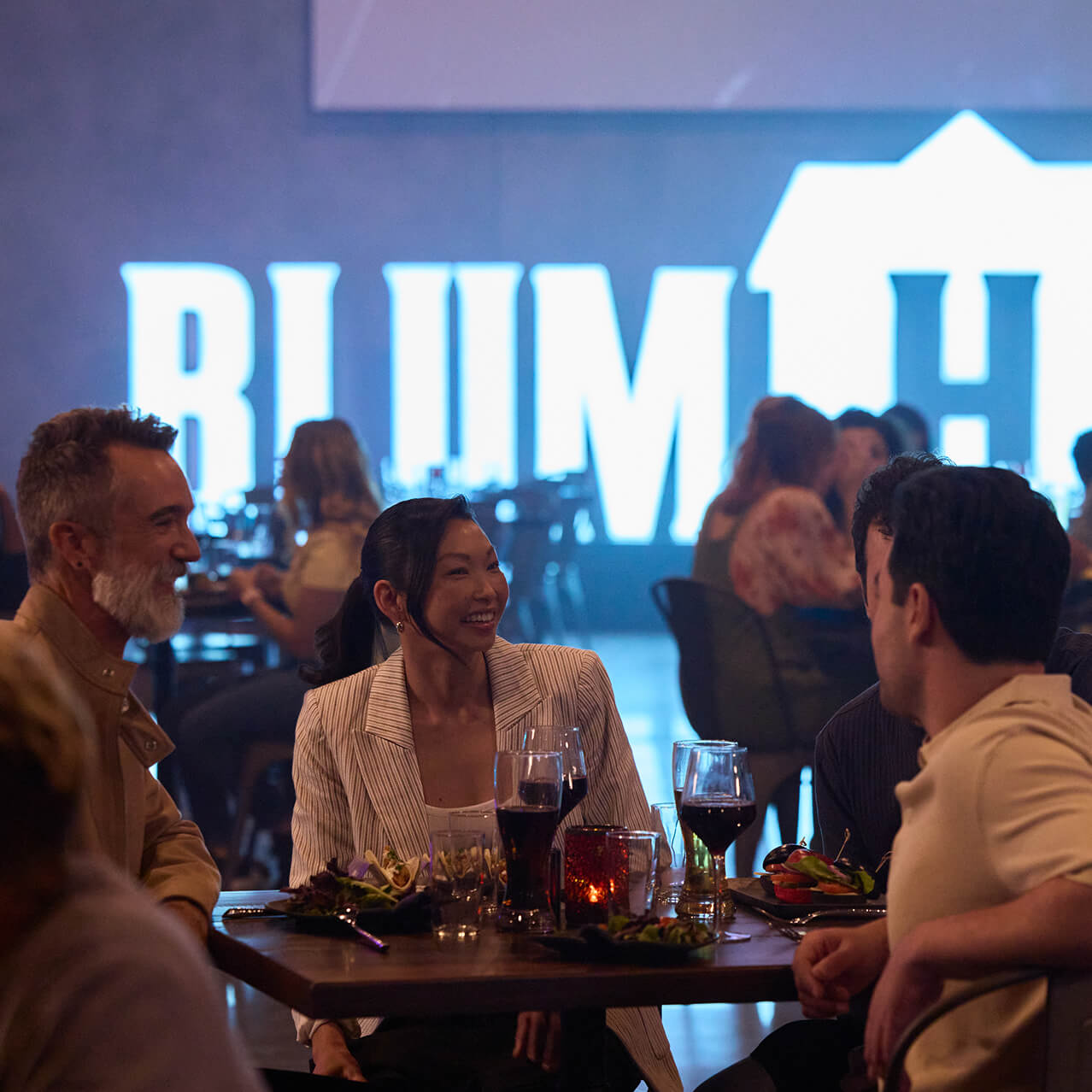Guests seated at a table enjoying food and drinks in front of a large illuminated Blumhouse sign.
