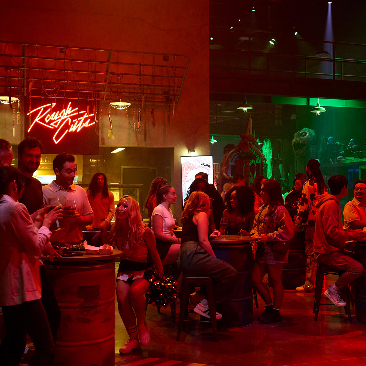 Guests gather around tables and drinks at a bar with a neon “Roughcuts” sign and dramatic red lighting.