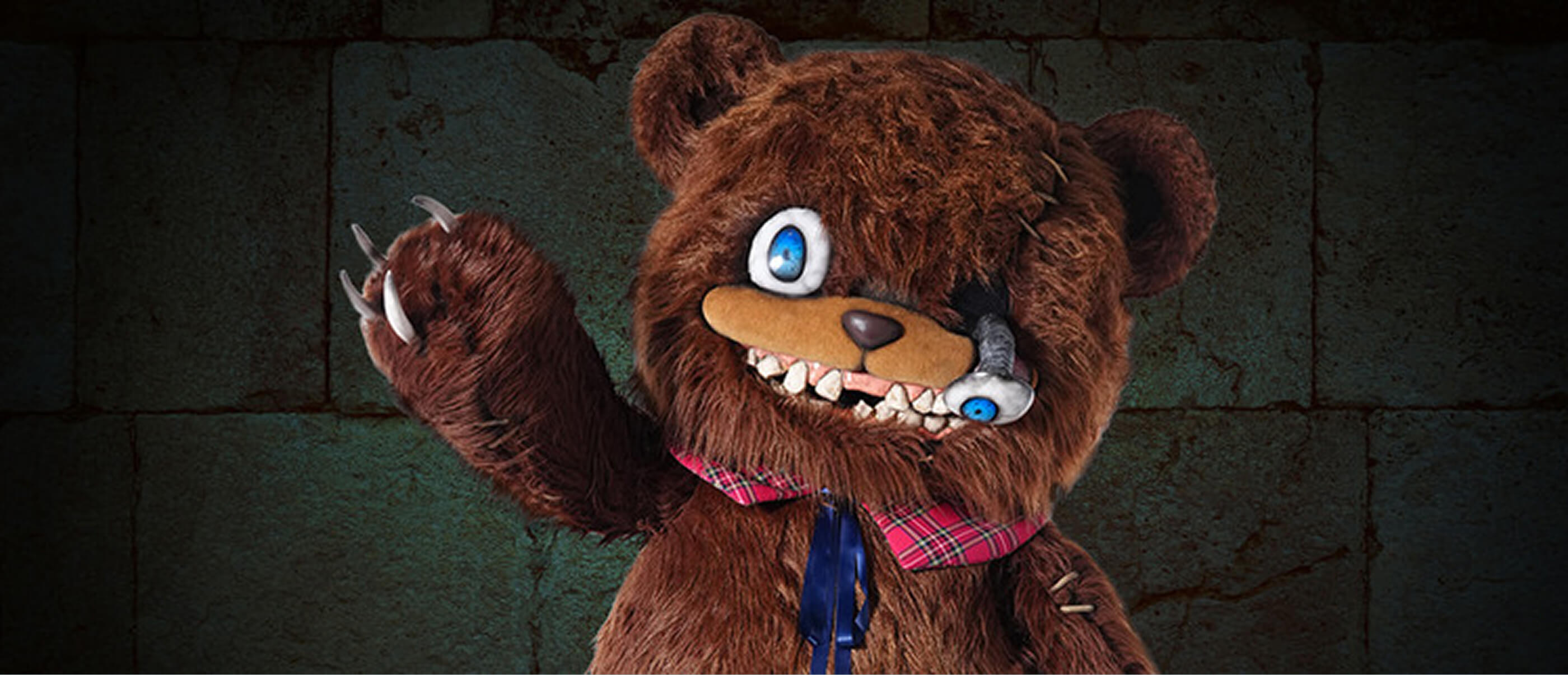A photo of the HamiKuma depicted as large smiling teddy bear with an eye falling out. He is waving while standing in front of a brick wall.