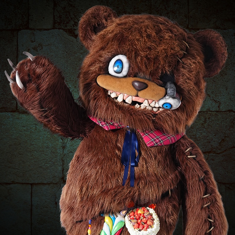 A photo of the HamiKuma depicted as a large smiling teddy bear with an eye falling out. He is waving while standing in front of a brick wall. 