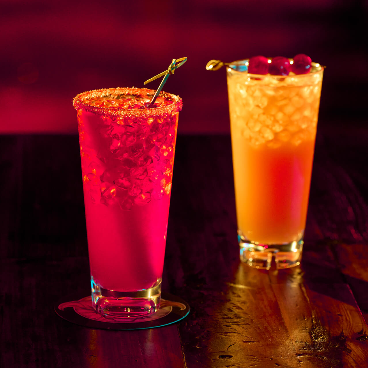 Two colorful cocktails in tall glasses on a bar, one bright red with a sugared rim and one orange with cherries.