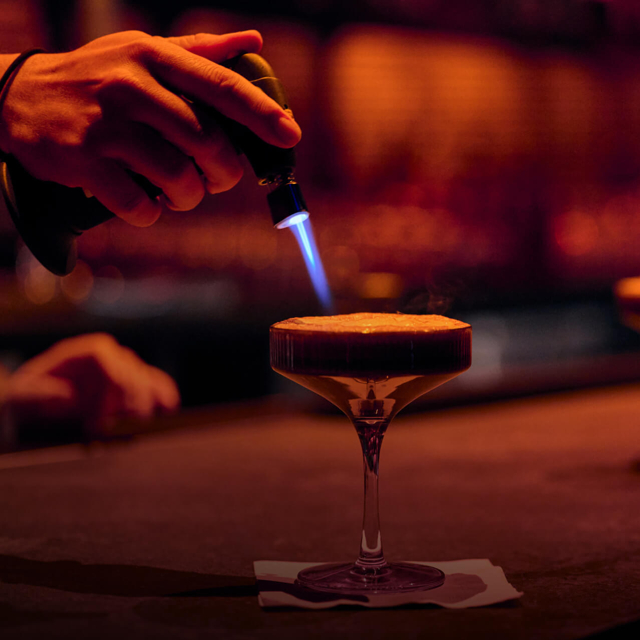 Bartender torching the foam on an espresso martini at a dimly lit bar.