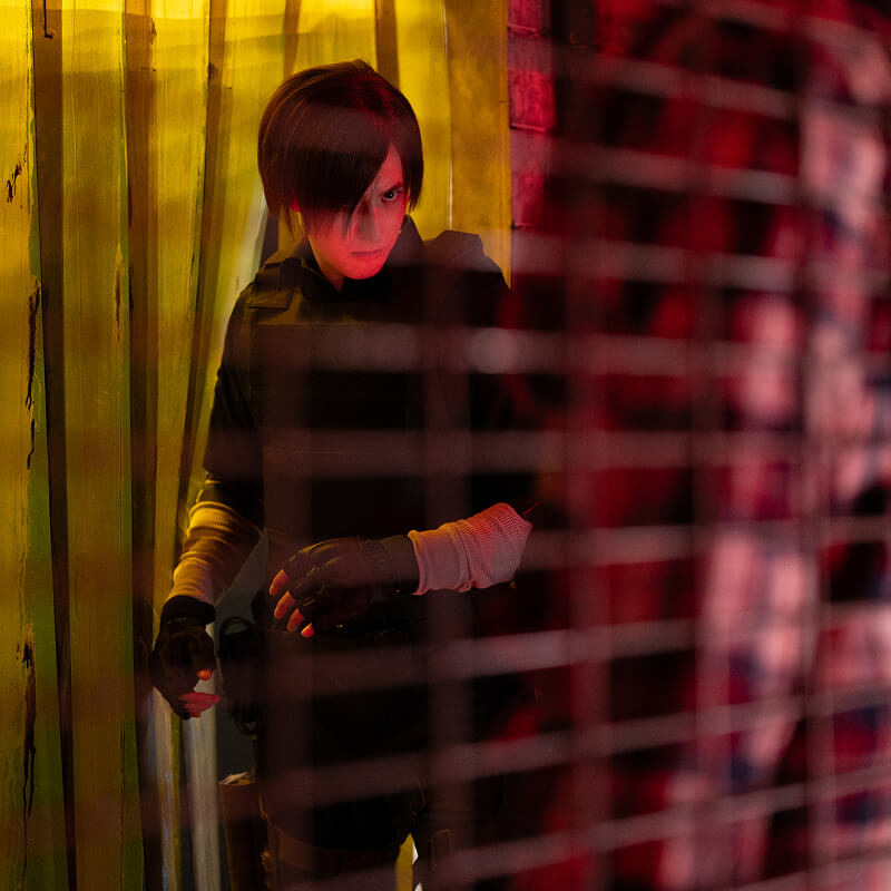 A photo of an actor portraying Leon S. Kennedy braces himself while watching a zombie behind a metal fence.