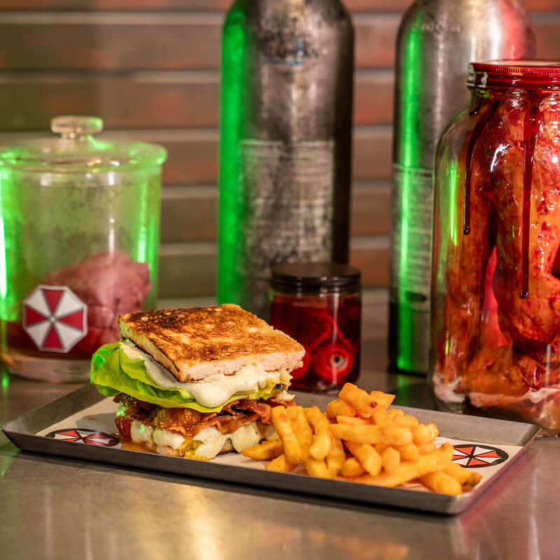 Photo of a meal consisting of a large toasted bacon sandwich, paired with fries and surrounded by bloody jars and containers.