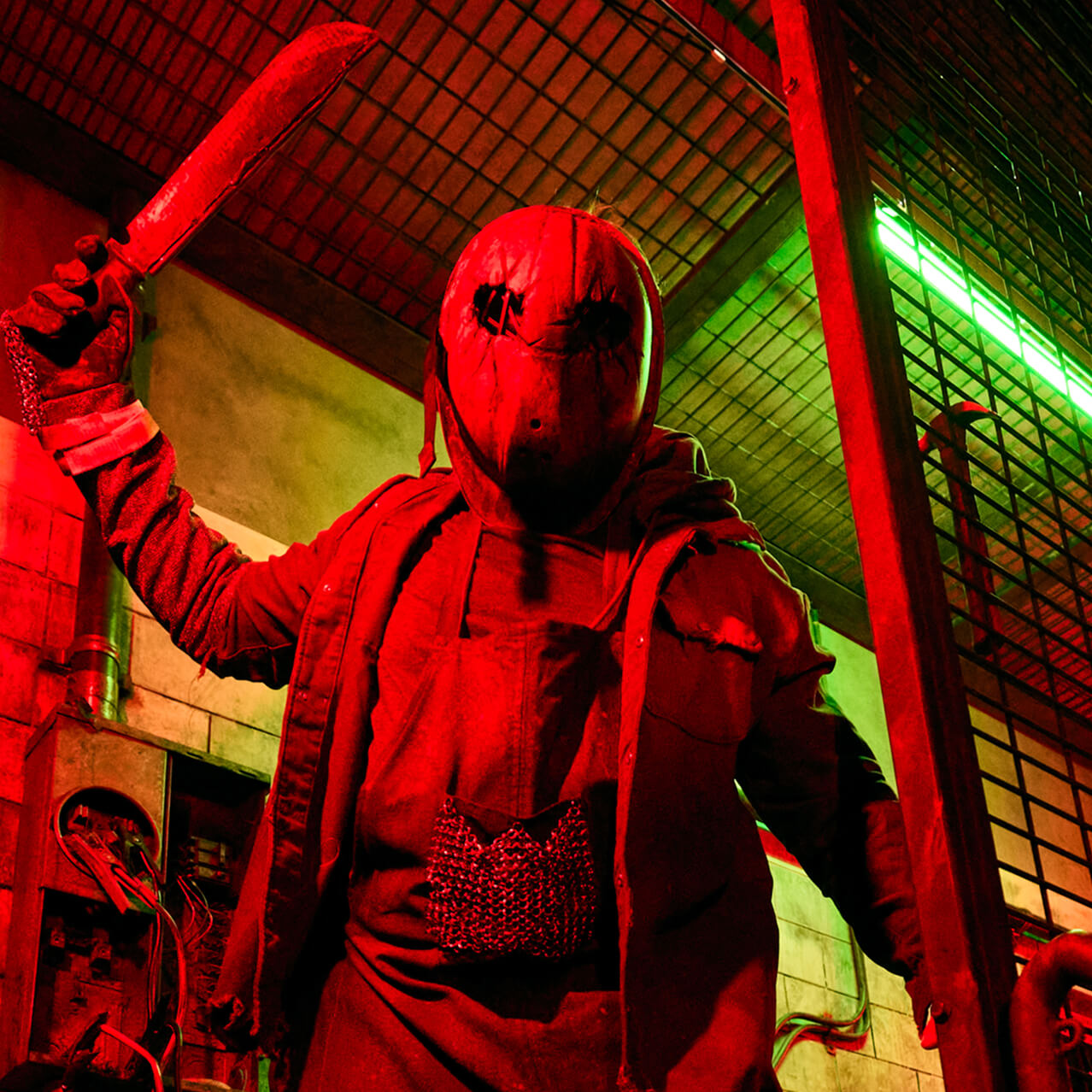 Masked scare actor holding a cleaver in a caged industrial room lit with red and green lights.