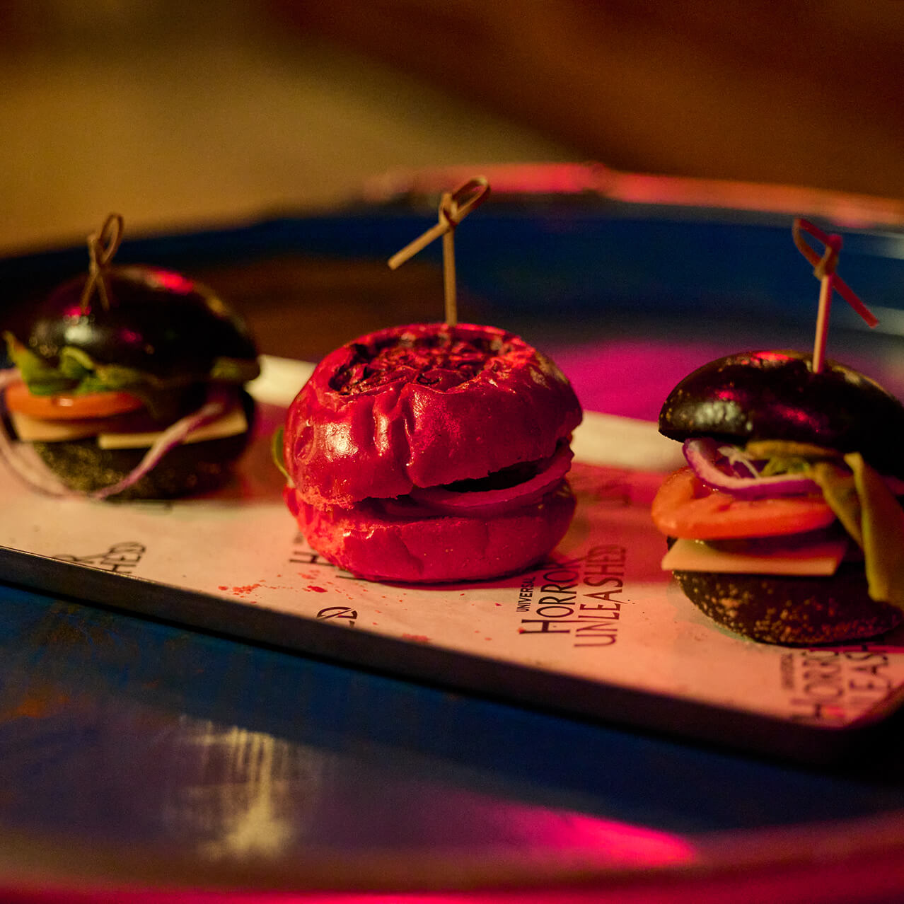 Three slider burgers on a tray, including one with a bright red bun, under colorful bar lighting.