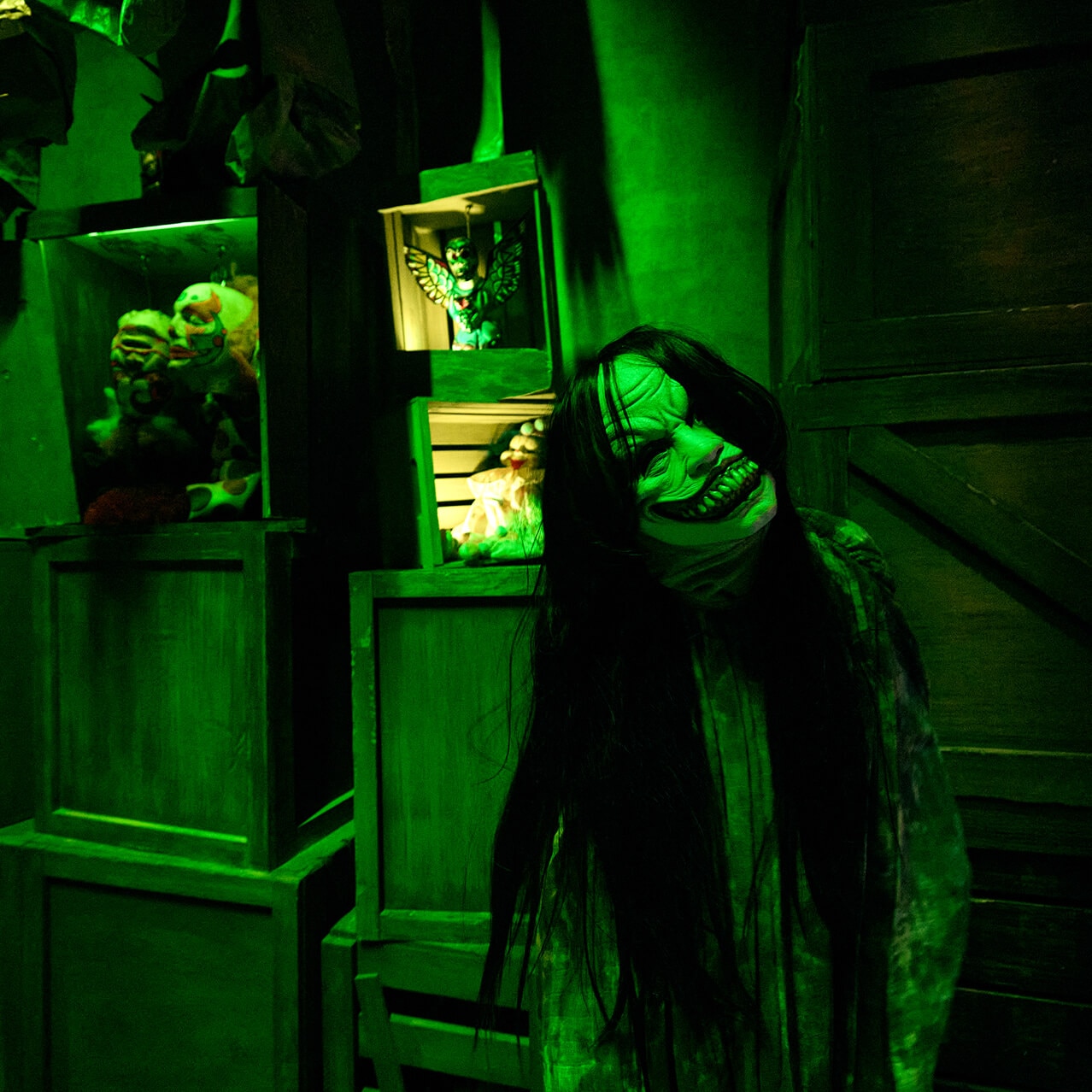 Long-haired scare actor wearing a twisted smiling mask inside a creepy prop warehouse filled with dolls.