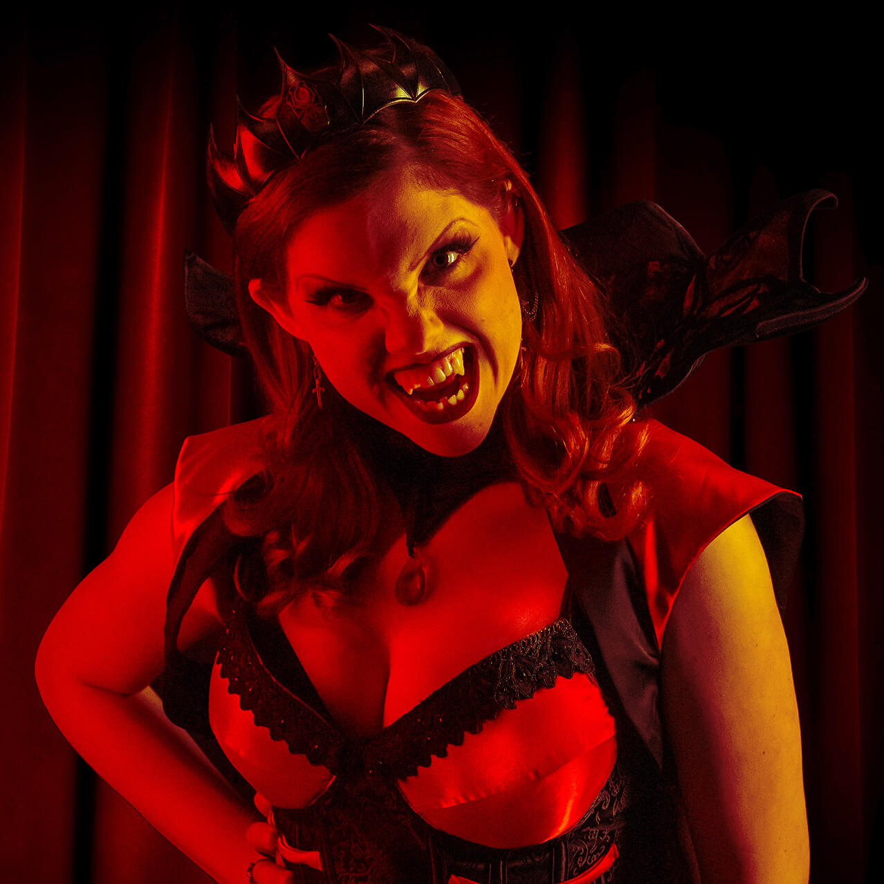 Vampire queen scare actor baring fangs, wearing a crown under red lighting.