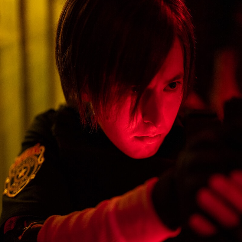 An actor portraying Leon S. Kennedy illuminated by red light, staring intensely during a tense moment.