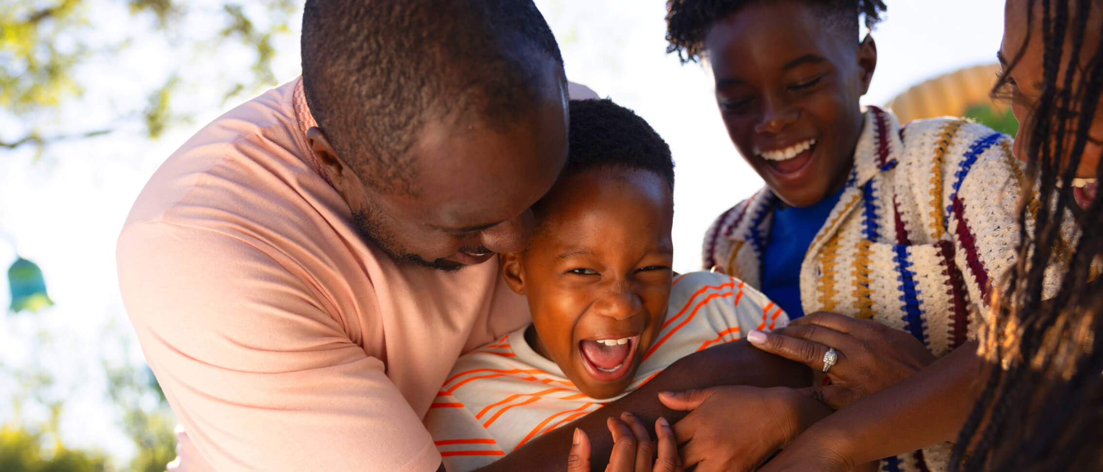 A smiling father playfully hugs and tickles his young son while his older son laughs beside them outdoors.