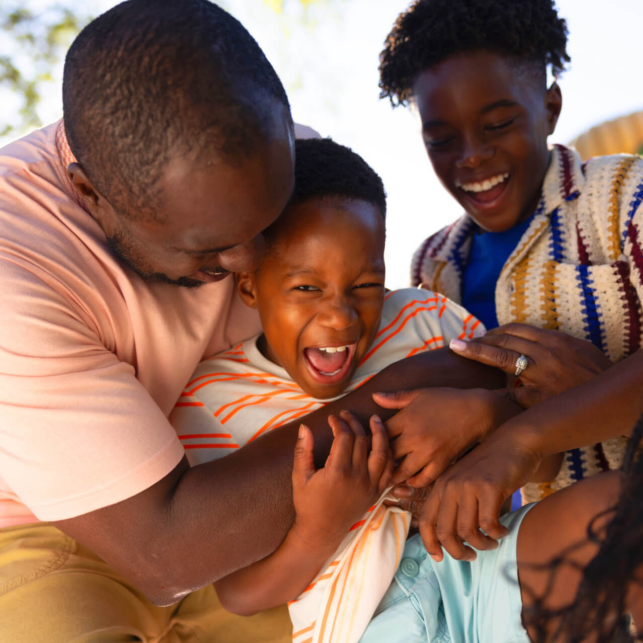A smiling father playfully hugs and tickles his young son while his older son laughs beside them outdoors.