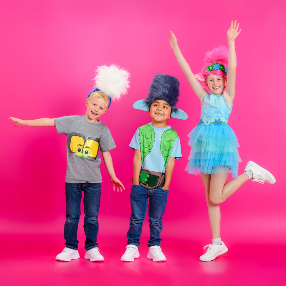 Three children dressed in Trolls-inspired costumes pose beside a Become Trollified graphic.