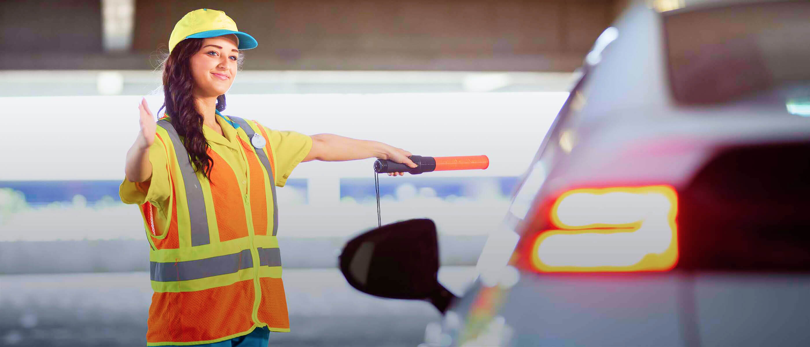 Parking garage attendant wearing reflective safety vest directing a car with illuminated traffic wand.