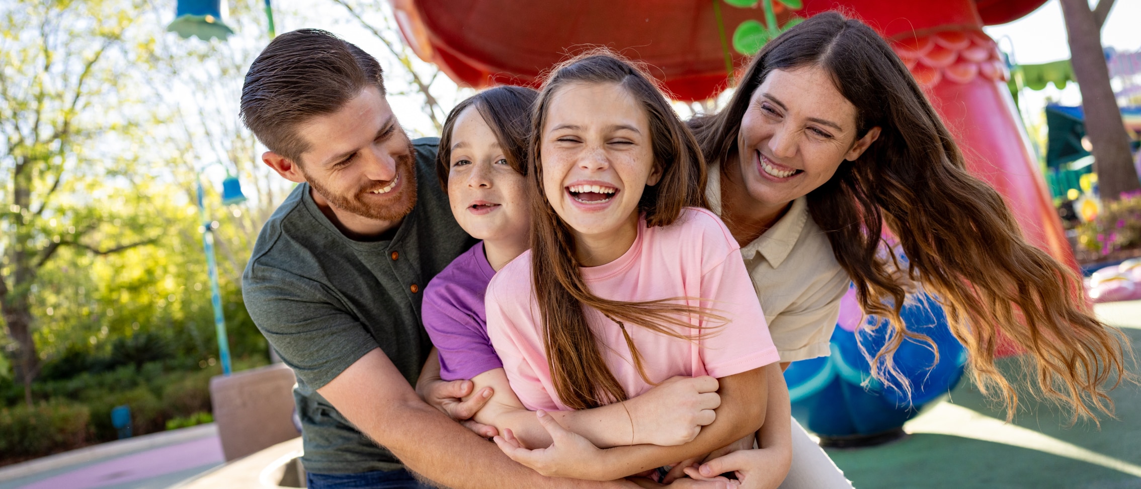 A smiling family of four hugging in front of a theme park ride.