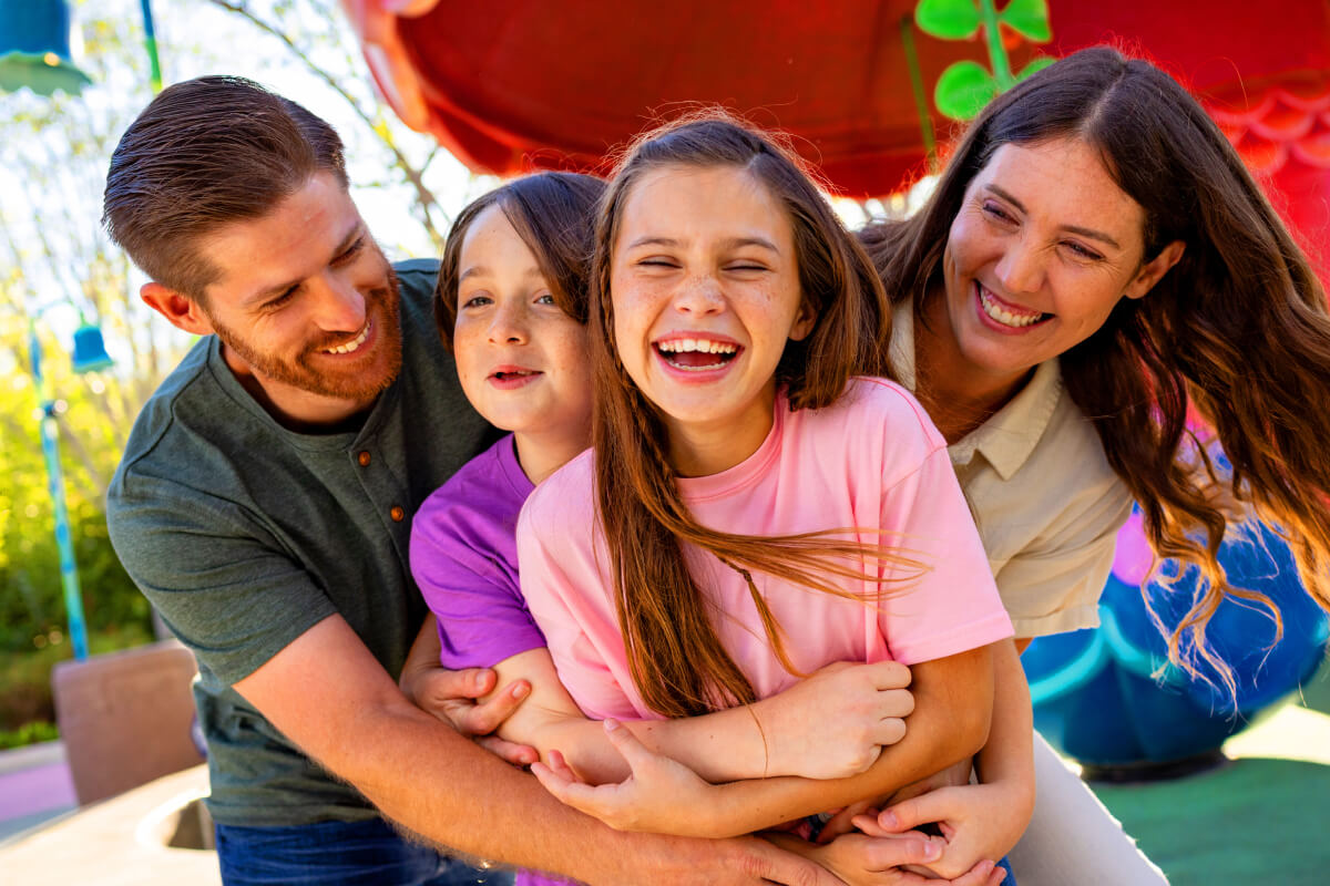 Two parents and their two young children smiling and hugging together outdoors at a colorful play area, with bright play structures in the background.