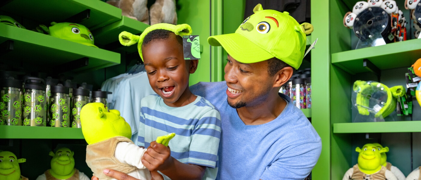 Un padre y un hijo jugando con un muñeco de Shrek mientras llevan gorros de Shrek en una tienda.