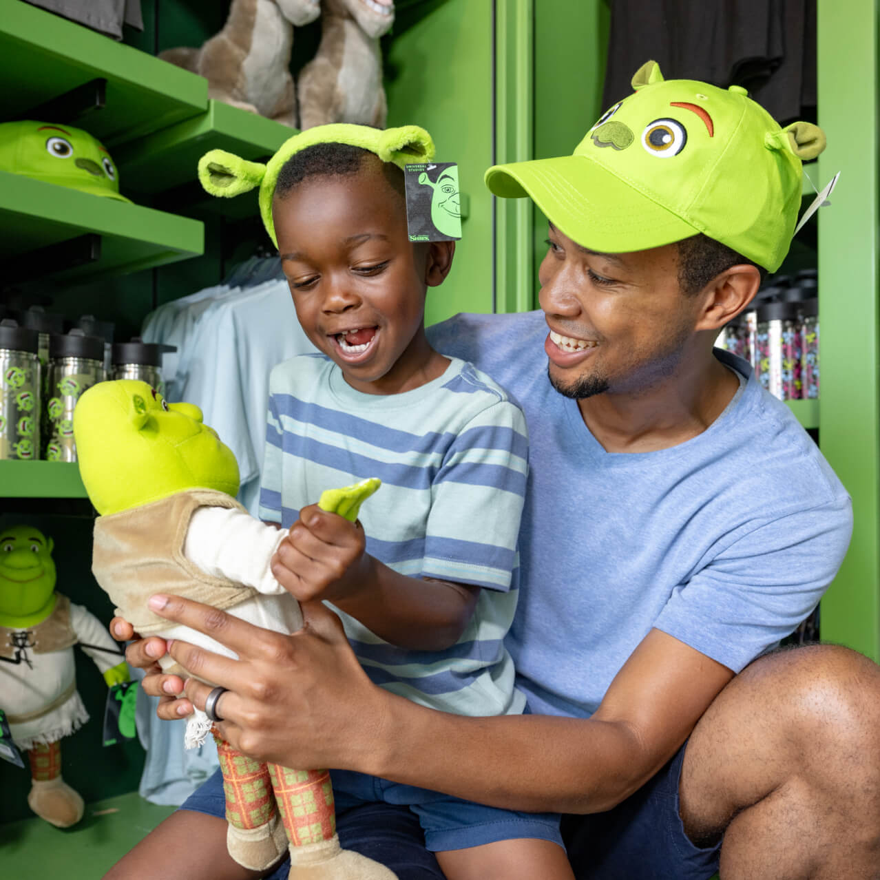 A father and son playing with a Shrek doll while wearing Shrek headwear in a shop.