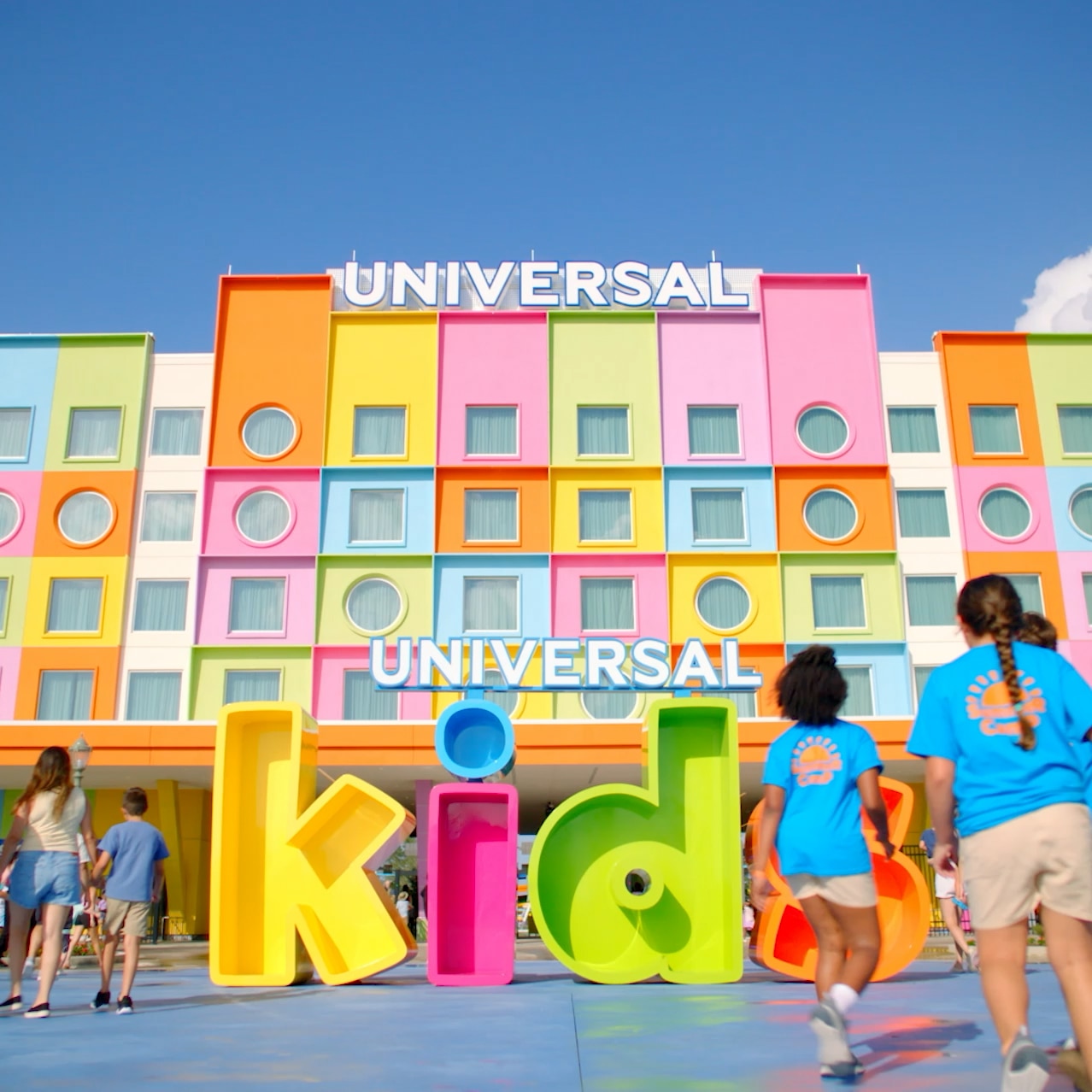 Families and kids walk toward the entrance of Universal Kids Resort, passing a large colorful “kids” sign in front of the bright and colorful Universal Kids Resort Hotel.