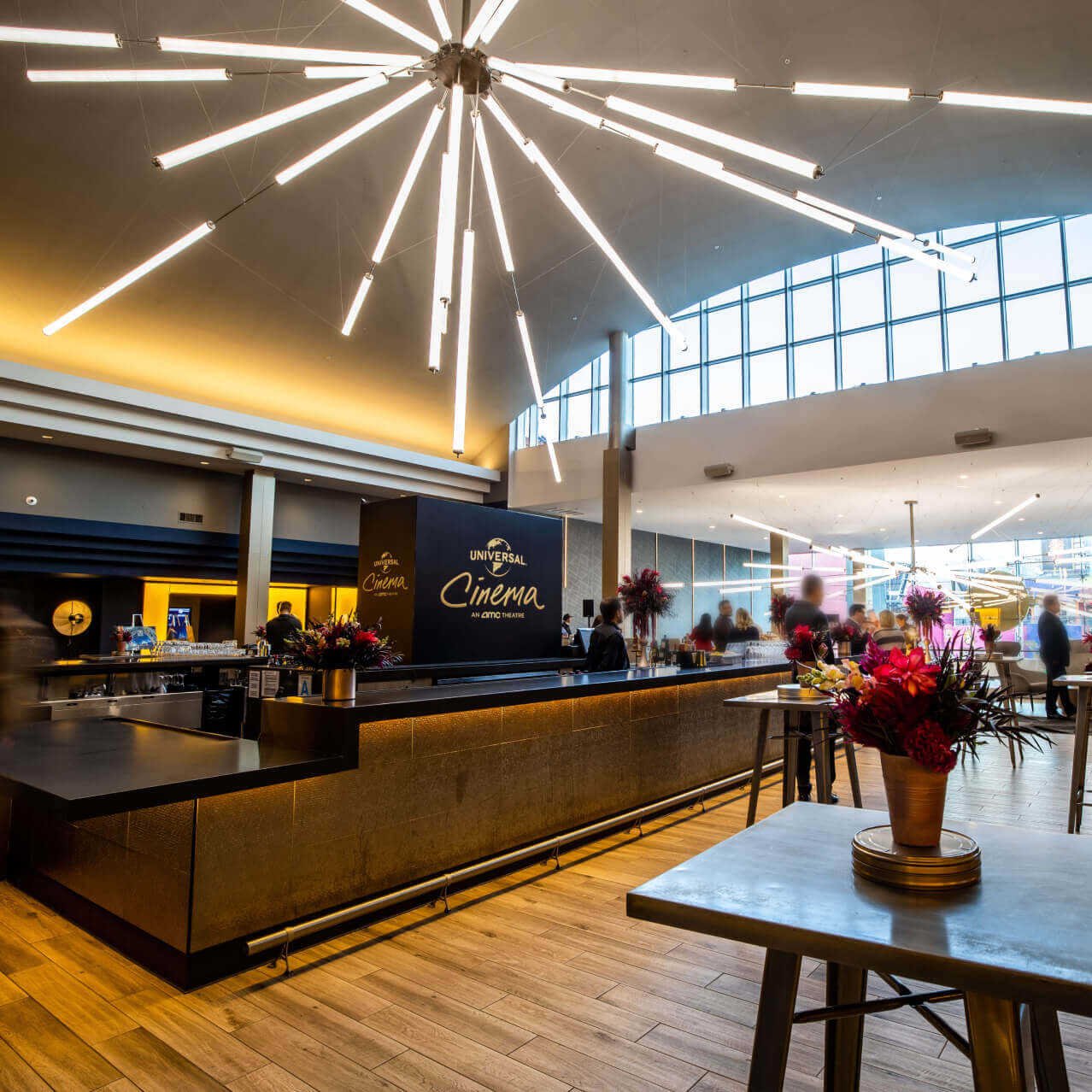 Flowers set up on tall tables in a bar area with wood floors, bright lights, large windows and a “UNIVERSAL Cinema” logo.