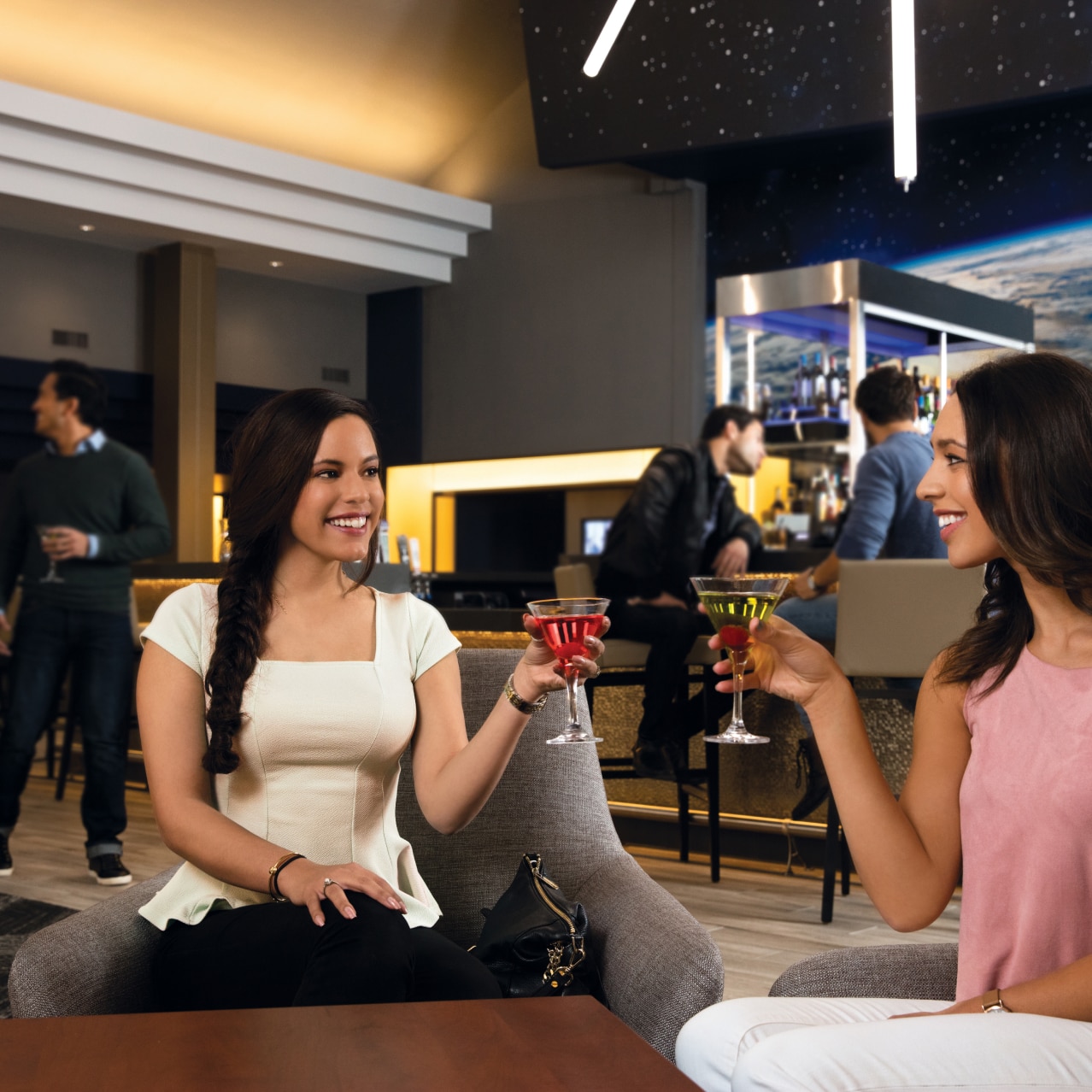 Two friends in armchairs smiling and holding cocktails with a bar in the background.