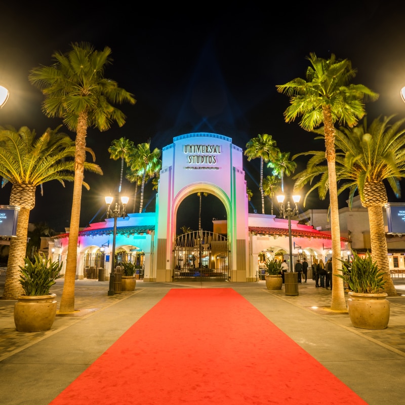 Red carpet lined with tables and floral arrangements leading up to the arch as it's lit with multicolored lights at the entrance to Universal Studios Hollywood.
