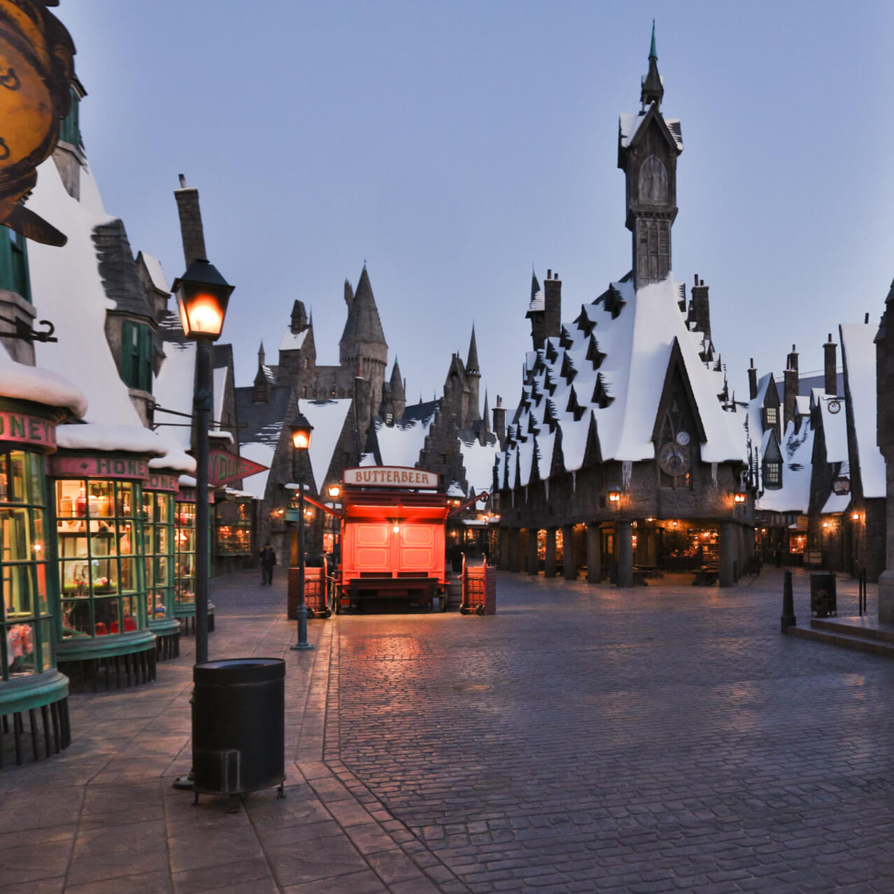 Snow-covered Hogsmeade™ village at dusk in The Wizarding World of Harry Potter™, with glowing lanterns, cobblestone streets, and themed storefronts.