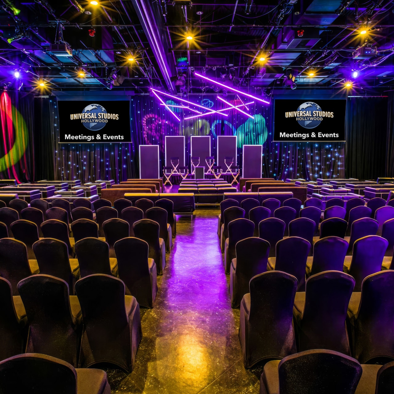 Dozens of chairs lined up in front of two large screens displaying the Universal Studios Hollywood logo above the words 'Meetings & Events" with colorful lights hanging above.