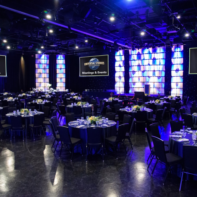 A venue set up for an event with dozens of tables and three screens in the background featuring the Universal Studios Hollywood Logo above the words Meetings & Events.