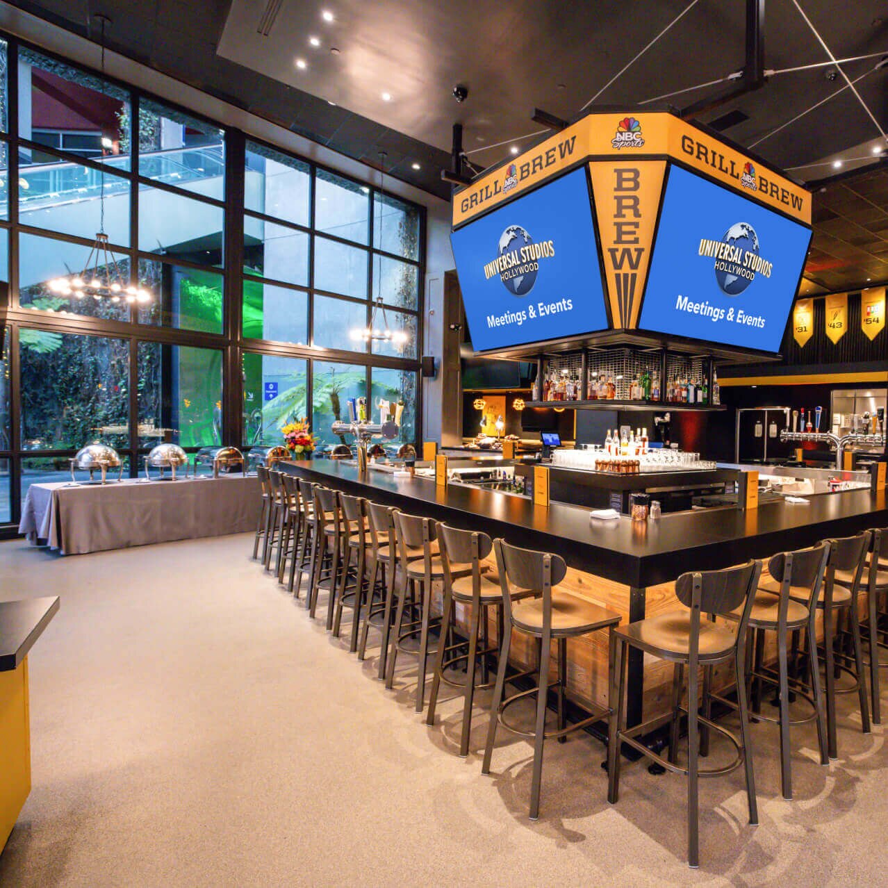 A square bar with high-top chairs and a large “NBC Sports GRILL BREW” sign by dishes and flowers set up on gray tablecloth.