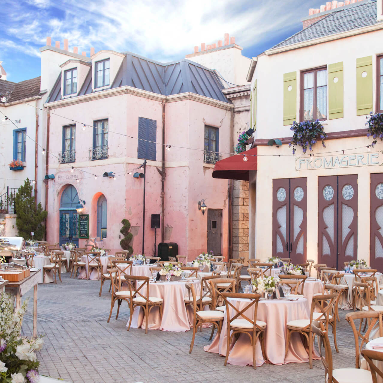 Elegant outdoor event setup on a European-style street set, with floral centerpieces, café lighting, and banquet tables.