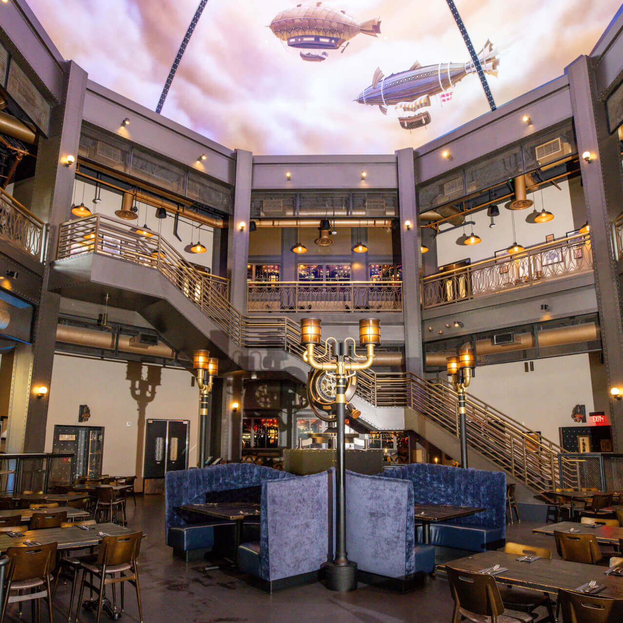 Varied seating including plush booths in an ornate, warmly lit restaurant with steampunk-style pipes and a staircase.