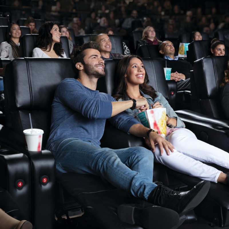 Two theatergoers in recliners with their feet up sharing a popcorn and watching a movie.