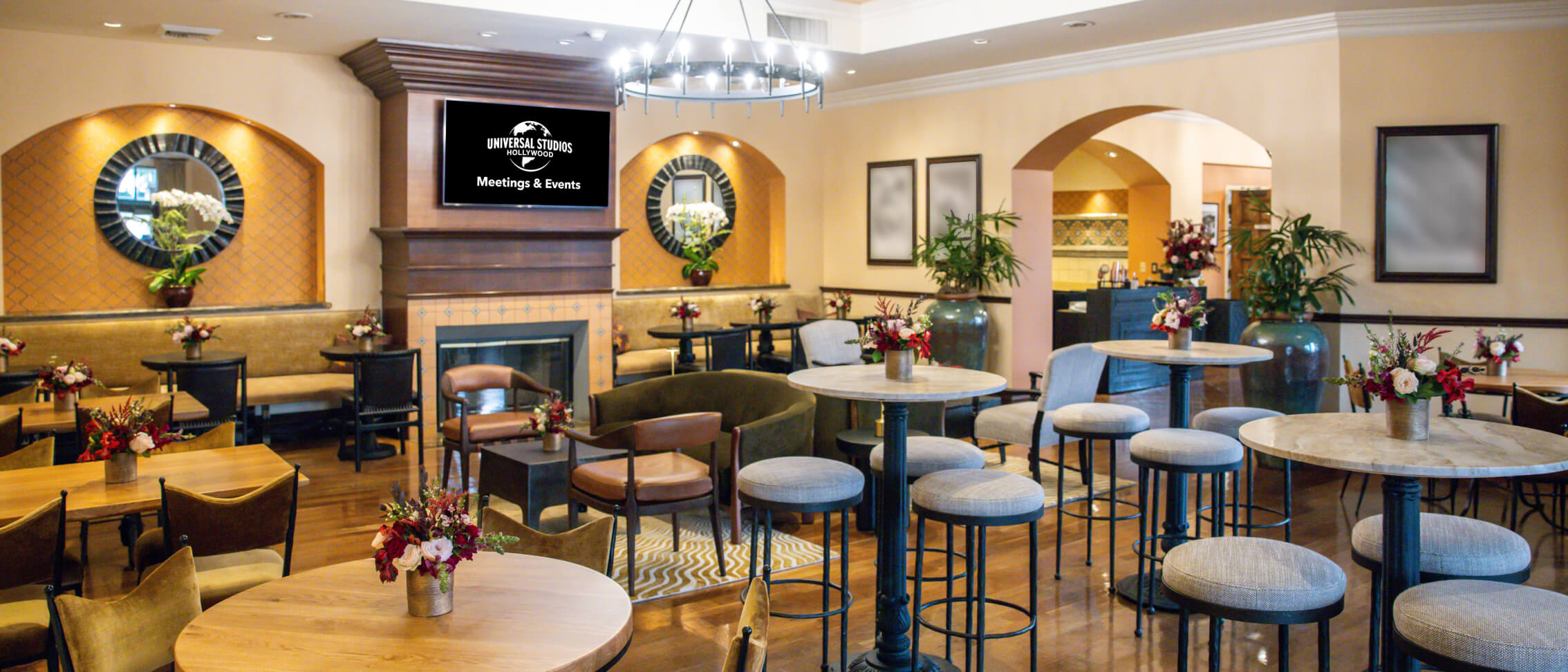 A warmly lit room with a variety of floral centerpiece tables and “Meetings & Events” on a screen mounted over a fireplace.