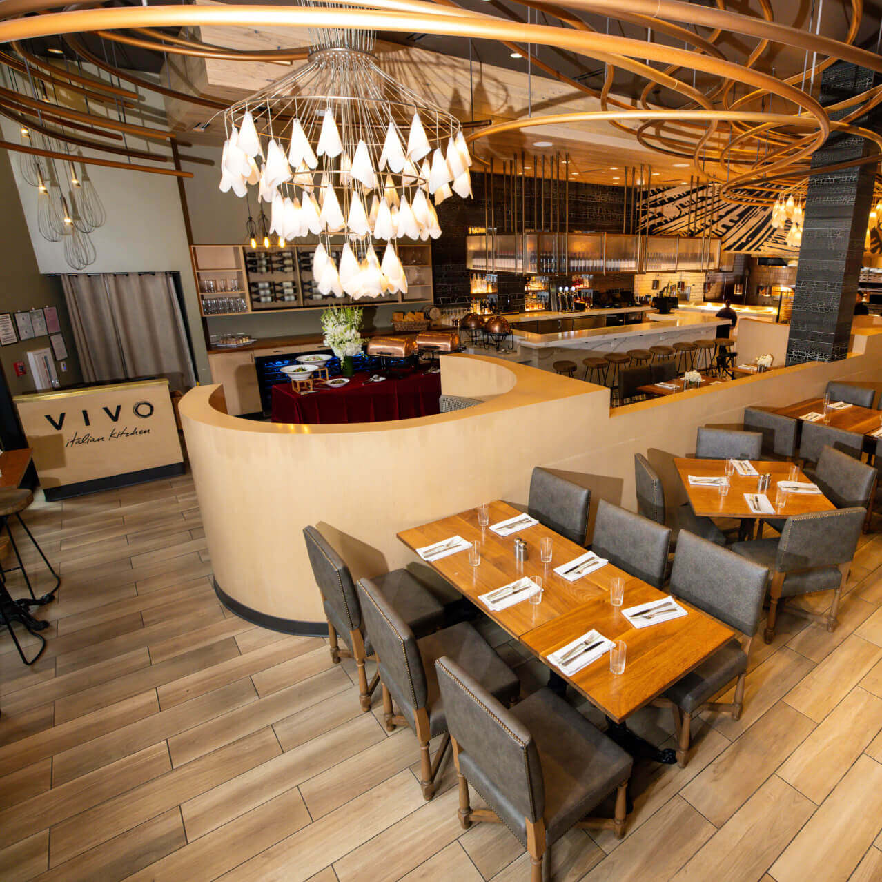 Table seating in a restaurant with wood floors, artsy light fixtures and ceiling accents, and a “VIVO Italian Kitchen” logo.