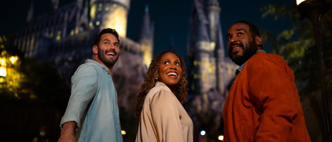 Three people turning around to smile with Hogwarts Castle lit up at night in the background.