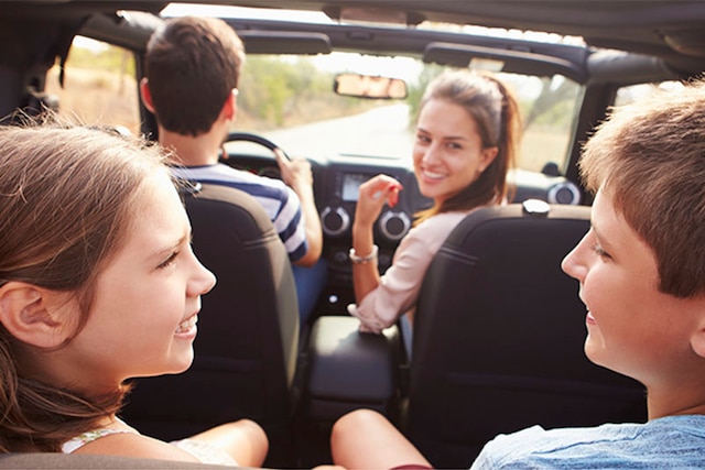 A happy family of four takes a drive in their rental car on a sunny day.