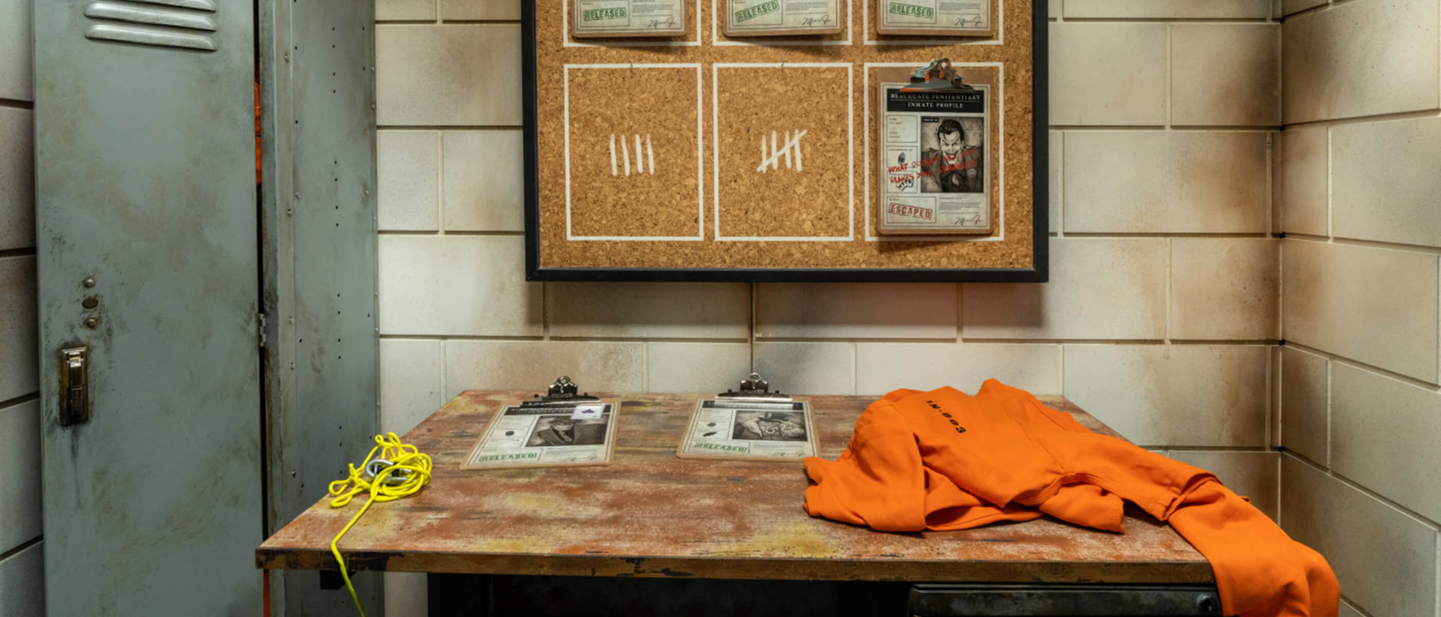 A jail-themed room containing a gray locker, bulletin board with a photo of the Joker, and a wooden desk with clipboards, an orange jumpsuit and a yellow rope.