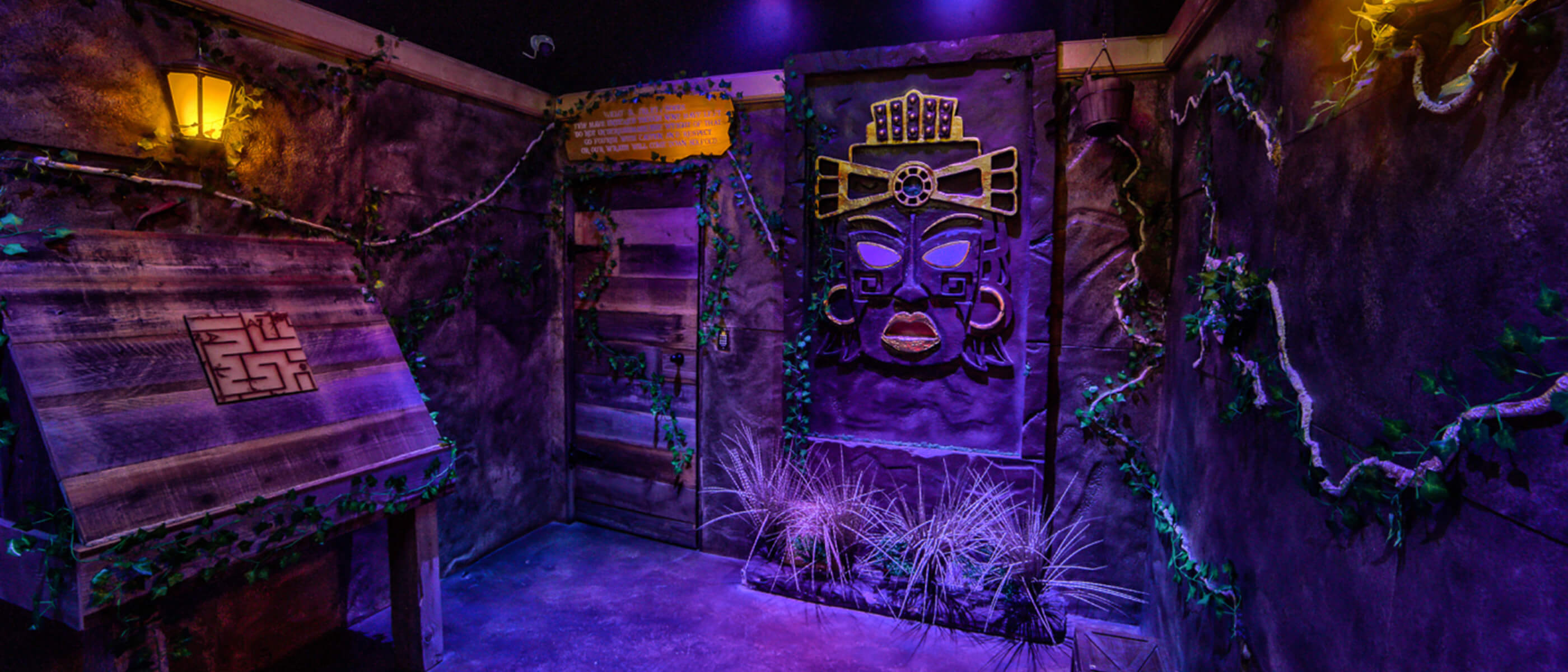 A dark purple-lit room with greenery on the walls and tribal art with yellow accent lighting.