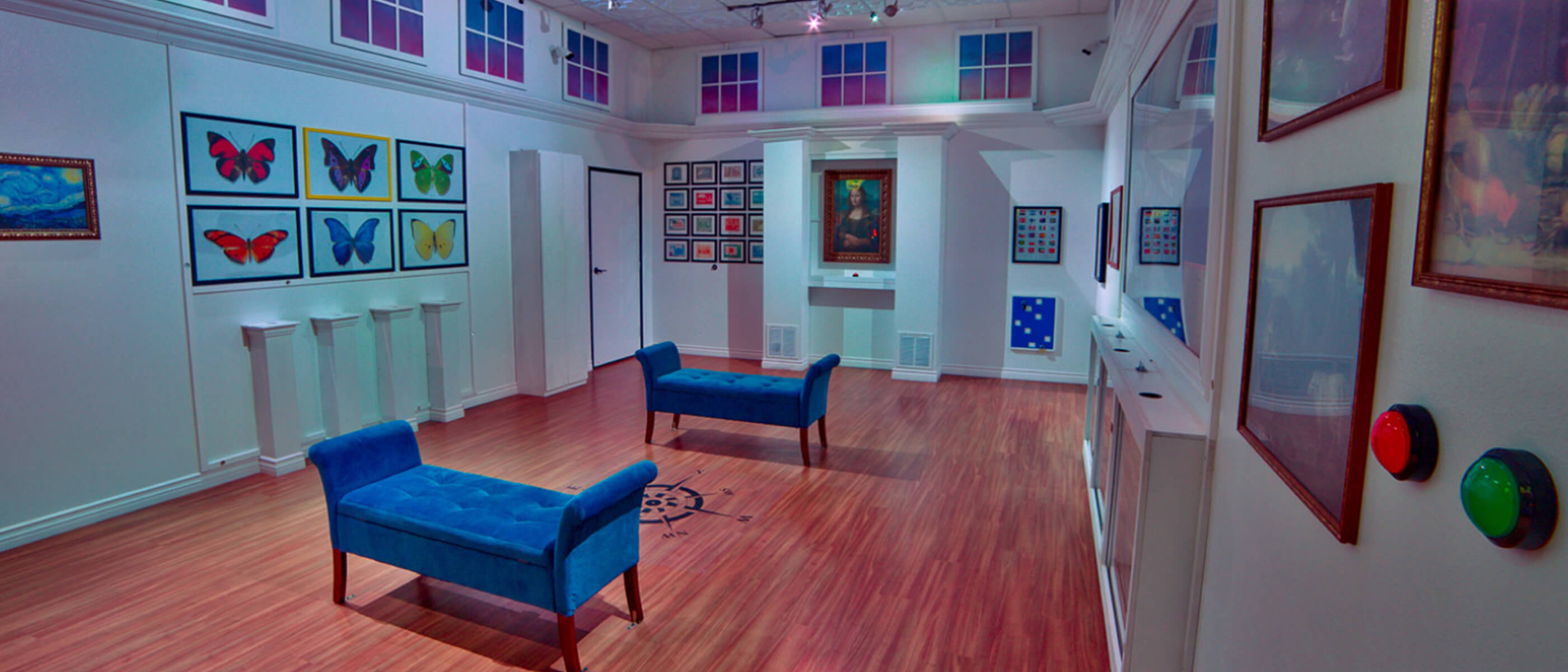 An art-gallery themed room with white walls and various types of art on the walls, with wooden floors and 2 blue seating fixtures in the middle.