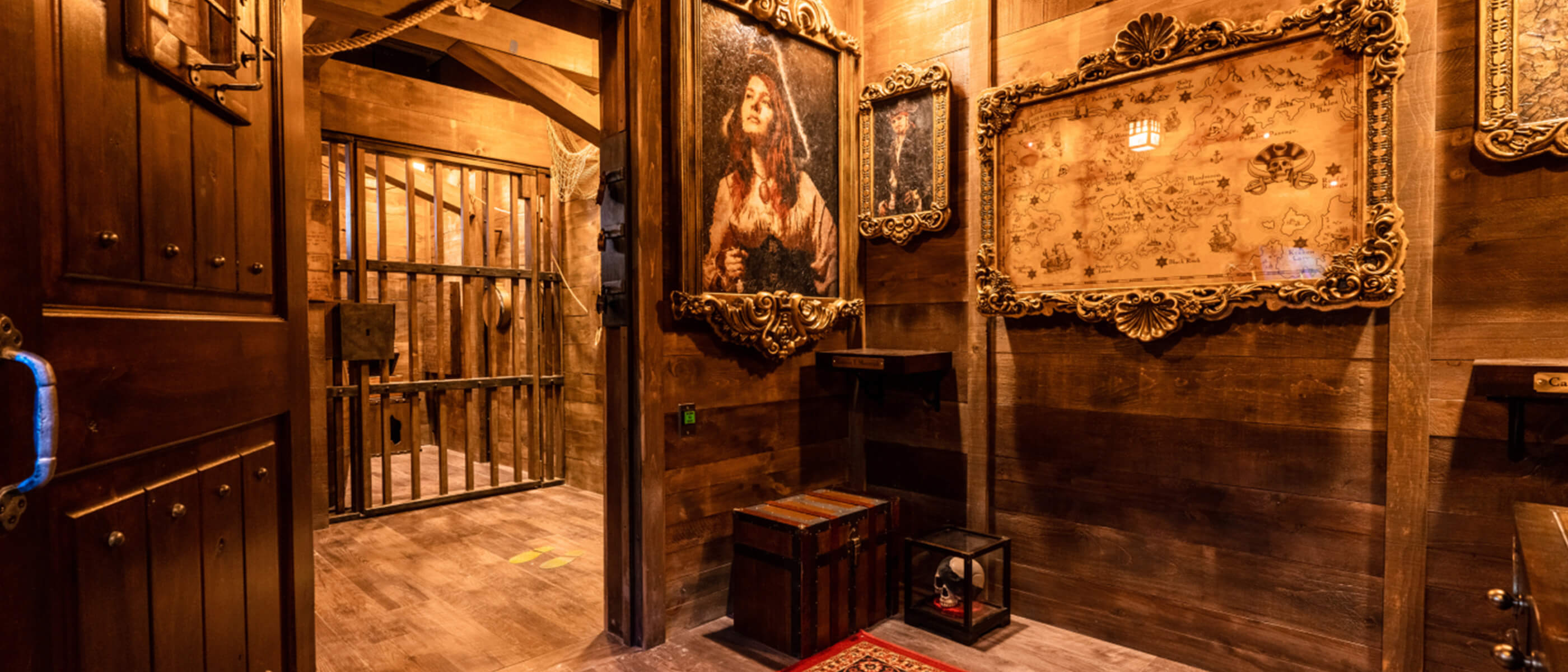 A room with wooden floors and walls and an open door to a jail cell, with pirate-themed art and a map on the walls.