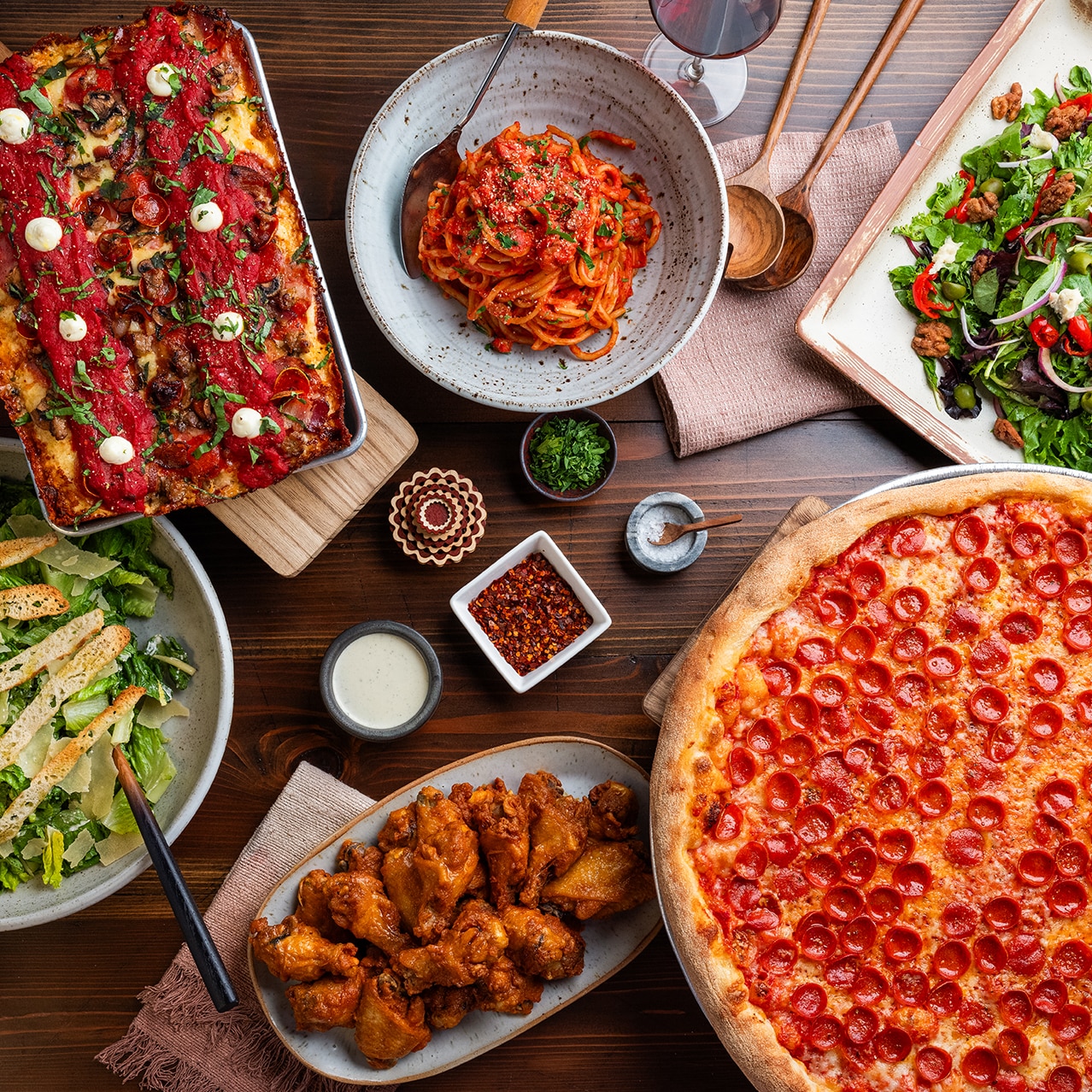 A tabletop featuring two pizzas, chicken wings, spaghetti, two salads and dipping sauces from Slice House.