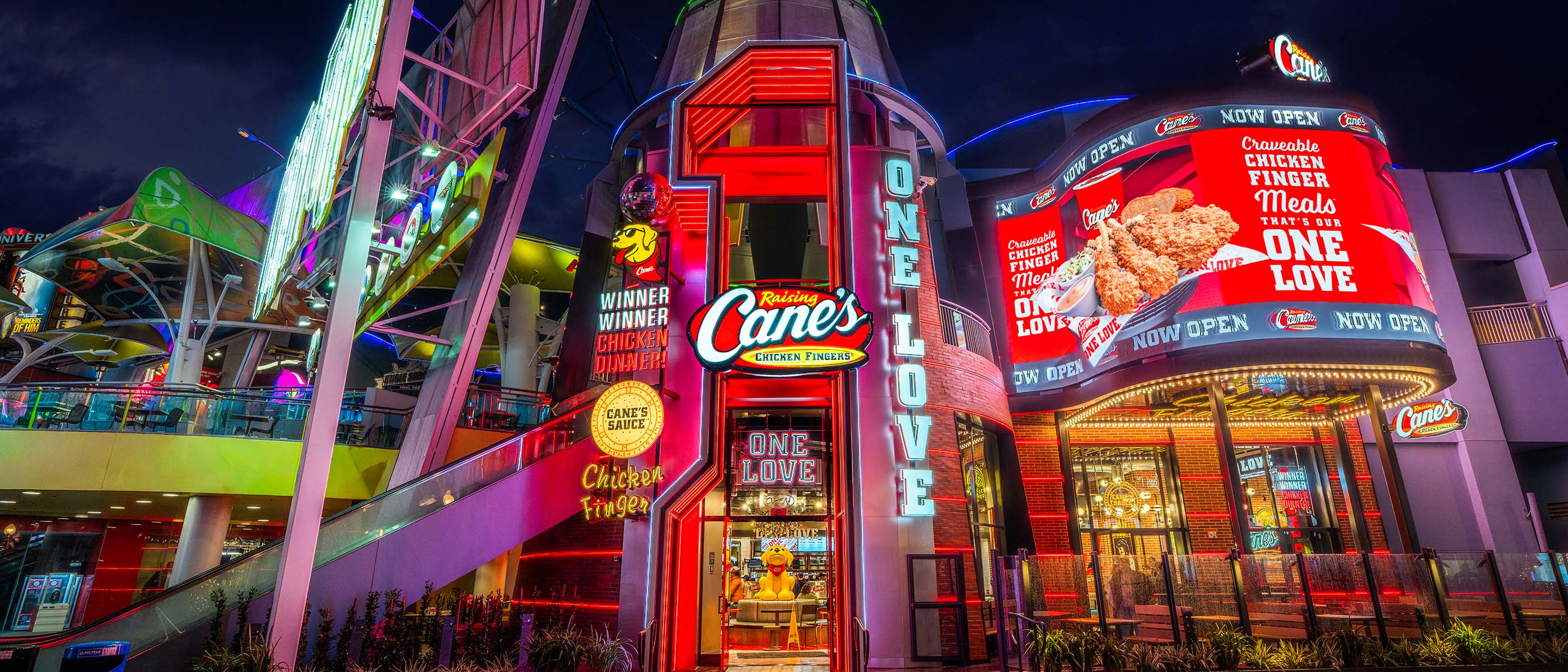 Exterior of “Raising Cane’s CHICKEN FINGERS” at night with neon lights and signage including “ONE LOVE” and a mascot dog.