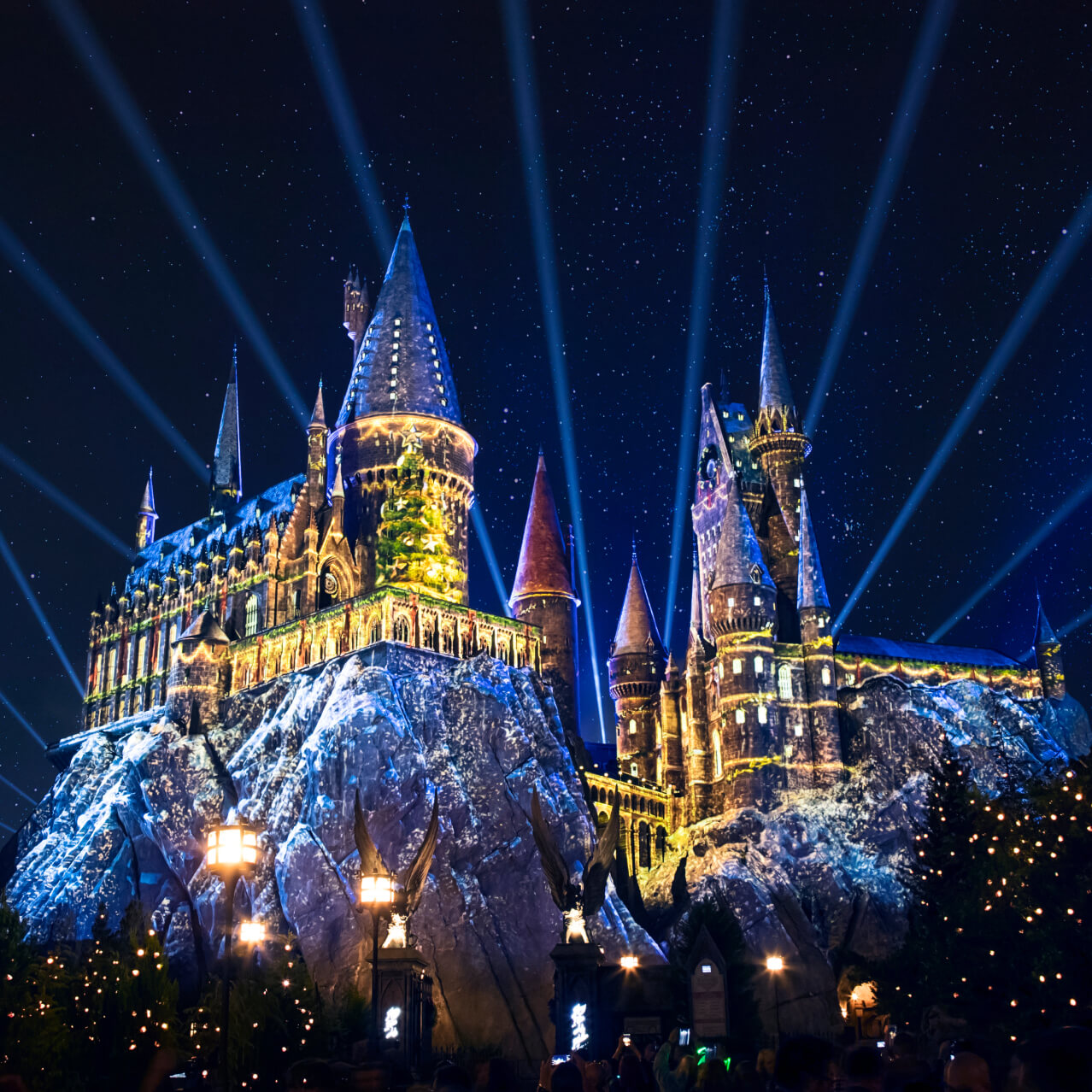 Hogwarts castle and the surrounding trees festively lit up in blue and yellow holiday lights with beams rising into the sky.