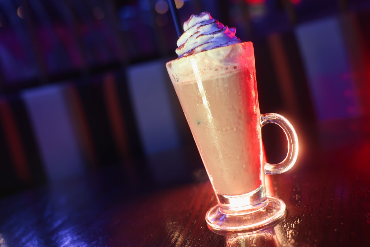A tall glass with a handle containing a coffee-colored drink topped with whipped cream and cinnamon on a darkened table.