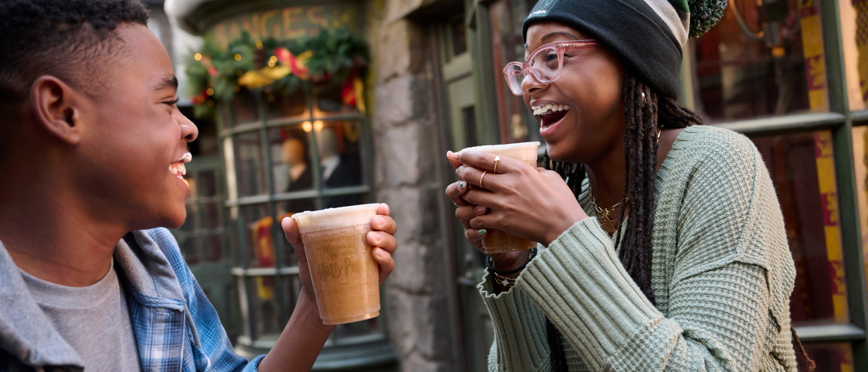 Friends laughing and holding cups of Butterbeer on the festively decorated streets of The Wizarding World of Harry Potter.