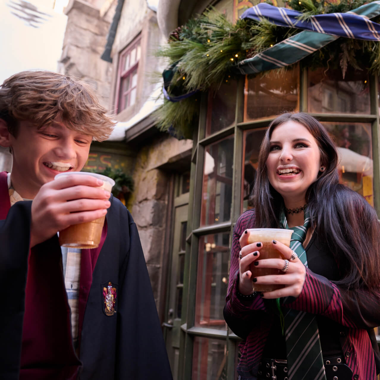 Two young Guests in wizarding gear laughing with foam mustaches as they enjoy Butterbeer by a festively decorated storefront.