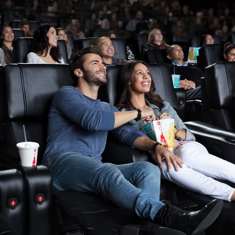 Two theatergoers in recliners with their feet up sharing a popcorn and watching a movie.